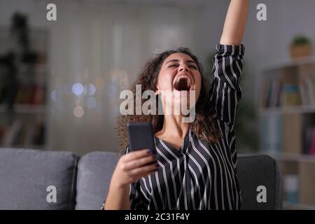 Femme élégante excité holding smart phone assis sur un canapé dans la nuit à la maison Banque D'Images