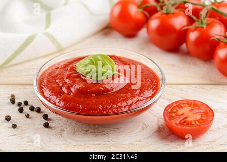 Ketchup de tomate épais et savoureux dans un bol en verre contre les tomates cerises rouges mûres sur une branche sur une table en bois blanc. Épices, assaisonnements et sauces. Gros plan. Banque D'Images