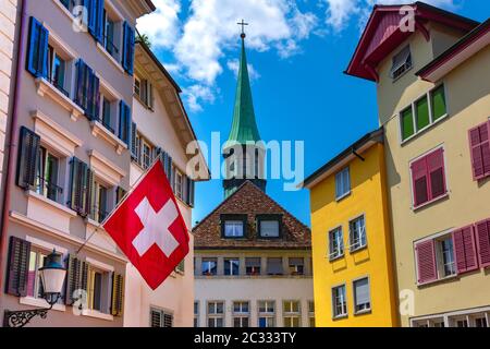 Drapeau suisse waving, église et façades colorées de maisons de la vieille ville de Zurich, la plus grande ville de Suisse en journée ensoleillée. Banque D'Images