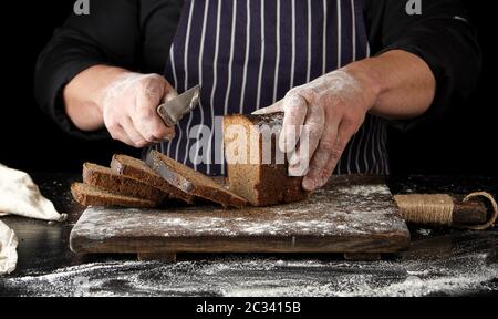 Chef en uniforme noir est titulaire d'un couteau de cuisine dans sa main et l'arrêt d'un des morceaux de pain cuit au four à pain de seigle marron sur une planche en bois Banque D'Images