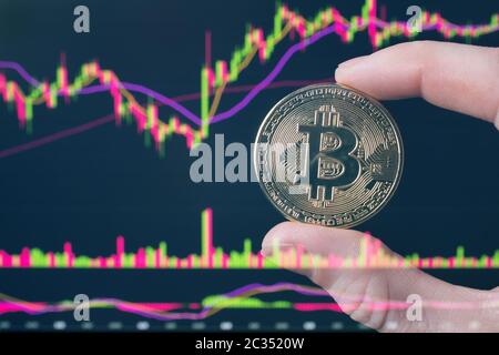 Tient une pièce Bitcoin assis devant un ordinateur portable analysant le tableau des prix d'un commerçant sur l'échange ou Forex. Bitcoin en main contre le fond de Banque D'Images