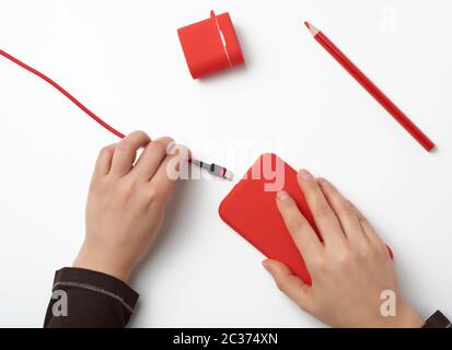 deux mains tenant un smartphone mobile rouge et un câble de chargement sur fond blanc, vue de dessus Banque D'Images
