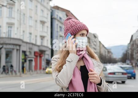 Femme préoccupée utilisant son téléphone portant un masque médical et des gants en ville Banque D'Images