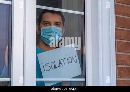 L'homme dans un masque médical regarde la caméra par une fenêtre. Auto-isolation dans la maison. Jeune homme attrayant dans des vêtements bleus avec masque médical Banque D'Images