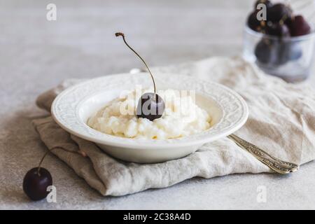 Un bol blanc avec un pudding au riz maison et une cerise foncée sur le dessus. Le bol est sur fond gris. Photo de concept. Banque D'Images