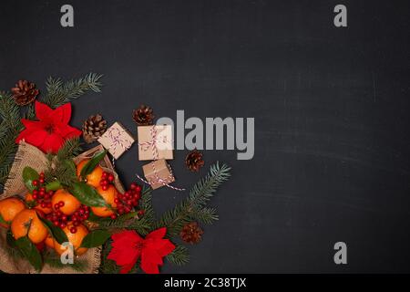 Vie de fête : baies et mandarines avec feuilles sur plateau en bois, fleurs rouges poinsettia, boîtes cadeaux et branches de sapin sur fond sombre. Haut Banque D'Images