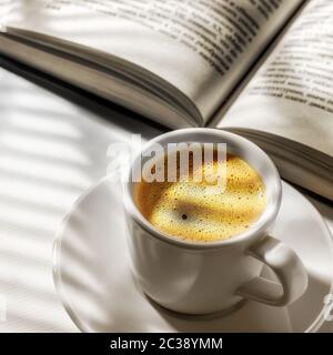 Une tasse de café noir et un livre ouvert sur une table blanche et l'ombre des stores