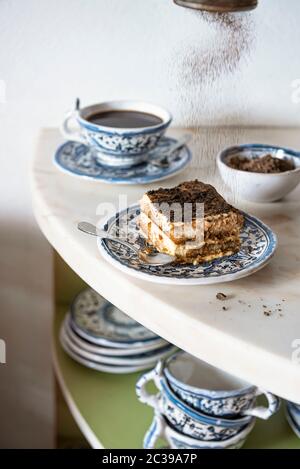 Tiramisu tranche sur une assiette bleue sur marbre avec café et poudre de chocolat tombant Banque D'Images