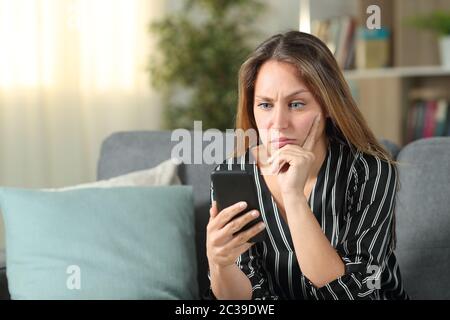 Confus woman checking mobile phone content assis sur un canapé dans la salle de séjour à la maison Banque D'Images