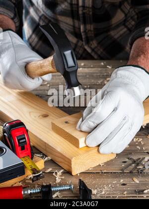 Close-up. Carpenter avec ses mains protégées par des gants, avec un marteau et des clous fixe une planche de bois. L'industrie de la construction, faire vous-même. Travail en bois Banque D'Images