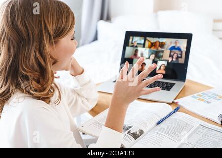 Photo de caucasienne bonne fille en agitant la main et en utilisant un ordinateur portable tout en étudiant en ligne à la maison Banque D'Images