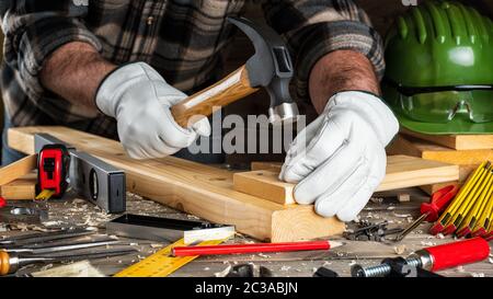 Close-up. Carpenter avec ses mains protégées par des gants, avec un marteau et des clous fixe une planche de bois. L'industrie de la construction, faire vous-même. Travail en bois Banque D'Images