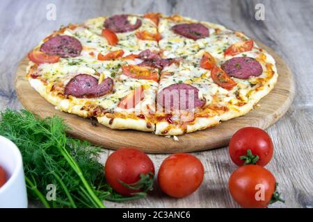 Pizza fraîchement cuite avec salami, champignons, herbes et plateau de fromage sur une table en bois près des tomates cerises, un bol de farine Banque D'Images