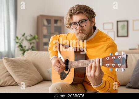 Jeune musicien masculin contemporain de vêtements décontractés jouant de la guitare acoustique tout en prenant le cours en ligne de faire de la musique à la maison Banque D'Images