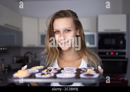 Jolie jeune femme qui fait cuire des cupkes sans gluten - en les sortant du four, en étant satisfaite du résultat Banque D'Images