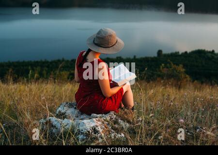 Une fille assise sur une pierre dans les montagnes lit un livre sur fond d'une grande rivière. Portrait au coucher du soleil. La fille dans le chapeau. Vacances à Banque D'Images