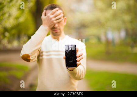 Beau jeune homme tient cassé écran de smartphone stressé de main sur la tête, choqué par la honte et le visage surprise, en colère et frustré. Peur et usse Banque D'Images