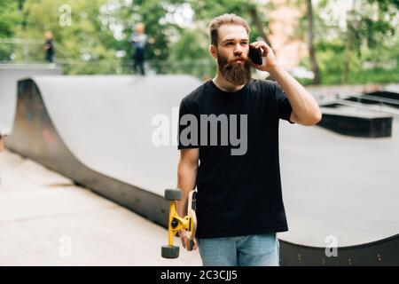 Un jeune homme avec un skateboard parle par un téléphone portable tout en étant assis dans un parc de skate par une journée ensoleillée. Banque D'Images