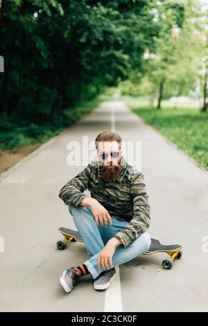 Jeune homme barbu longboarder dans des vêtements décontractés assis sur la longue planche ou le skateboard à l'extérieur. Urbain, sous-culture, concept de skateboard Banque D'Images