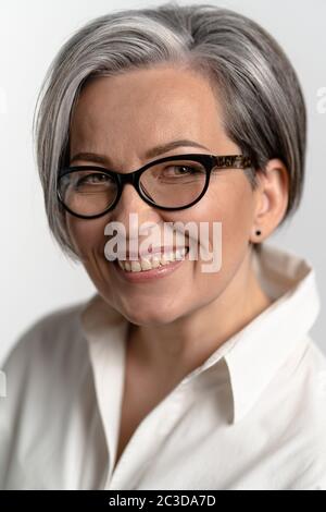 Jolie femme aux cheveux gris souriant à l'appareil photo. Femme mature en lunettes et chemise blanche sourit largement. Concept d'entreprise. Concept d'éducation. Gros plan Banque D'Images