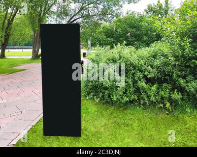 Un stand d'information rectangulaire en noir Uni avec un emplacement pour le texte se dresse sur une pelouse verte dans un parc d'été, sur un fond de buissons verts et Banque D'Images
