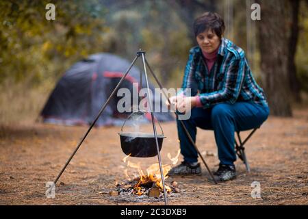 Femme âgée préparant des aliments sur feu de camp tout en vivant dans la vie sous tente dans la forêt. Banque D'Images