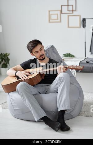 Beau jeune homme à la moustache noire assis dans un sac de haricots et jouant de la guitare à la maison pendant la quarantaine Banque D'Images