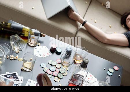 Vue au-dessus de l'homme ivre dormant sur un canapé avec ordinateur portable après la fête, la nourriture désordonnée et des verres sur la table avec des jetons de poker Banque D'Images