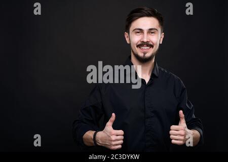 Portrait d'un jeune homme excité debout sur fond noir et montrant le pouce vers le haut comme symbole de qualité Banque D'Images
