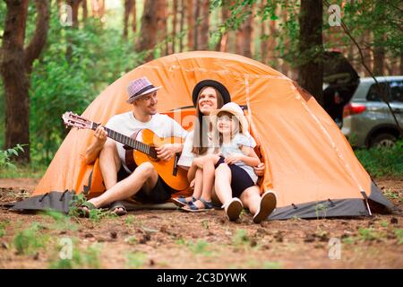 Portrait de beaux jeunes parents et de leur petite fille mignonne qui s'embrasse, regardant l'appareil photo et souriant pendant que l'homme joue de la guitare dans le camp forestier. HAP Banque D'Images