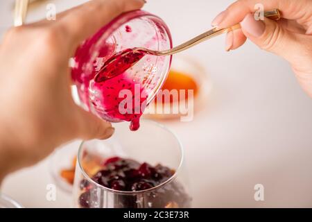 Main femelle avec cuillère à café mettant la confiture de framboise maison dans le verre tout en faisant du yaourt avec le miel et d'autres ingrédients Banque D'Images