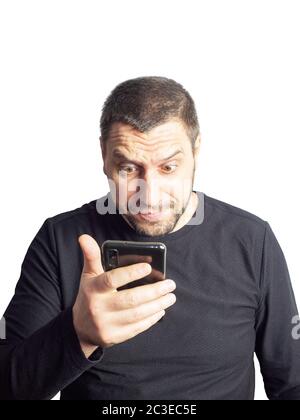 Un homme avec une barbe et dans un chandail noir regarde le téléphone avec ses sourcils relevés et ses yeux renflés. Banque D'Images