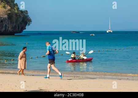 Railay, Thaïlande - 19 février 2019 : un homme court sur la plage le matin. Une femme marche le long de la plage de sable. Dans l'eau, un jeune couple nage en kayak. Banque D'Images