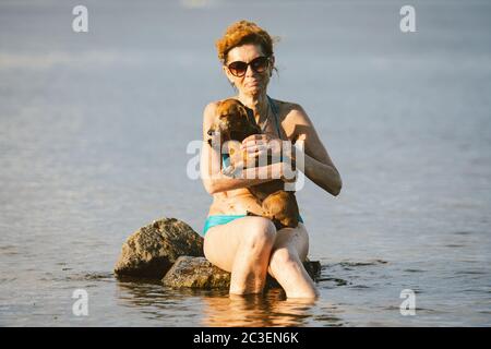 Un petit chien vieux brun de race Dachshund est assis sur les mains d'une femme mature dans un étang. Thème pour passer l'été à nager dans la rivière avec un animal de compagnie Banque D'Images