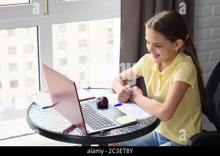 Fille regarde joyeusement l'écran de l'ordinateur portable tout en étant assis à une table dans l'appartement et faire des devoirs Banque D'Images