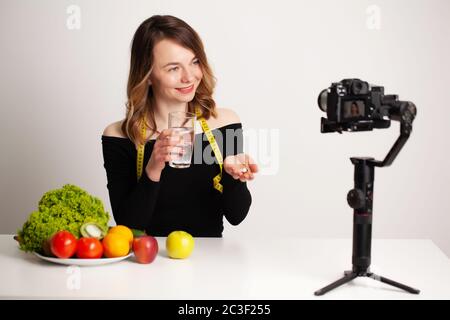 Femme tenant un comprimé de vitamine d'huile de poisson oméga-3 et un verre d'eau potable dans sa chambre et enregistrant un blog vidéo sur son téléphone. Banque D'Images