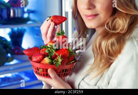 Portrait d'une belle femme blonde avec plaisir manger des fraises fraîches savoureuses près du réfrigérateur ouvert, bon choix, nourriture végétarienne, perte de poids Banque D'Images