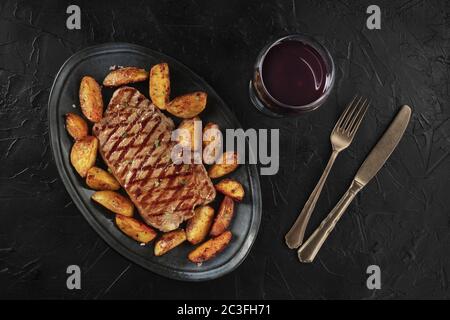 Steak grillé au bœuf avec pommes de terre cuites au four, avec un verre de vin, tiré du dessus sur fond noir Banque D'Images