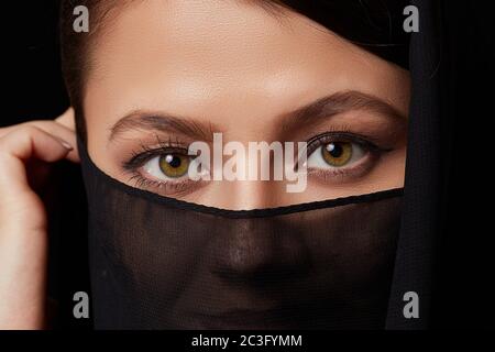 une jeune femme à poil brun en vêtements noirs avec sa tête couverte et son visage recouvert. sur fond noir isolé. Banque D'Images