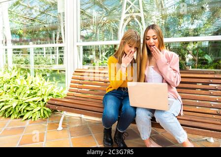 Deux jeunes filles caucasiennes attrayantes et expressives utilisent un téléphone portable et un ordinateur portable assis sur un banc en bois Banque D'Images