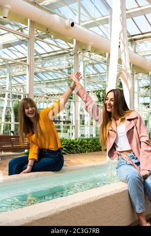 Deux jeunes filles caucasiennes attrayantes avec des cheveux blonds se secouer les mains avec leurs bras dans l'air en souriant à une fontaine à l'intérieur d'une pièce lumineuse avec le Banque D'Images