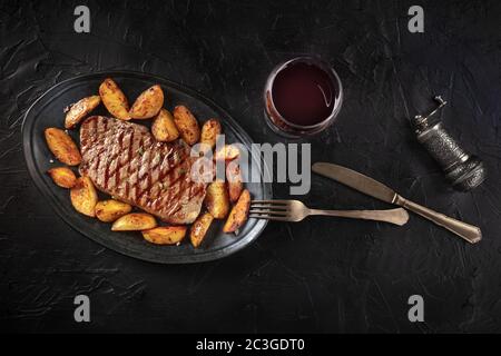 Steak de bœuf grillé avec des quartiers de pommes de terre cuits et du vin rouge, tiré du dessus sur fond noir Banque D'Images