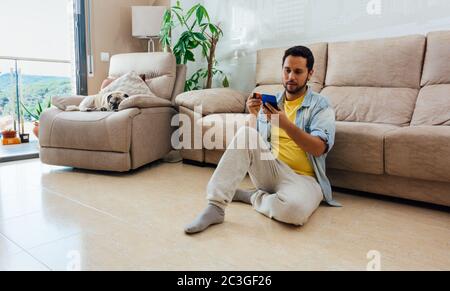 Photo grand format d'un homme assis sur le sol à la maison et en utilisant son téléphone Banque D'Images