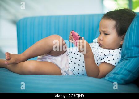 Bébé chubby utilisant un téléphone cellulaire rose par elle-même sur le canapé. Photo de haute qualité Banque D'Images