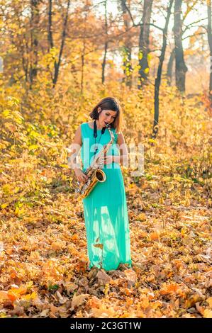 jolie fille avec cheveux noirs dans une robe bleue joue le saxophone alto au milieu du feuillage jaune dans un parc en automne Banque D'Images