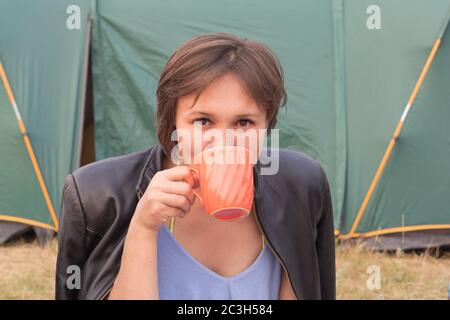 Jolie jeune femme dans un haut et une veste, boire du thé d'une grande tasse et regarder l'appareil photo sur le fond de la tente. Le tourisme domestique concep Banque D'Images