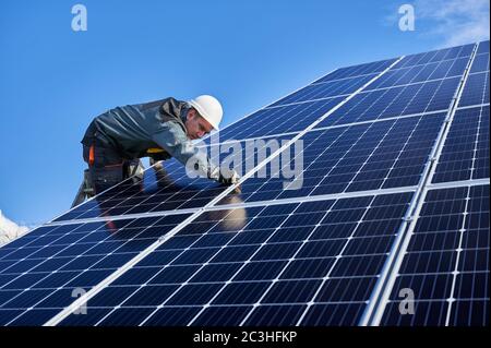 Belle et brillante surface de batterie solaire et homme travailleur portant un uniforme, debout sur l'échelle. Électricien mâle installant des modules solaires le jour ensoleillé, vue à angle bas. Concept d'énergie alternative Banque D'Images