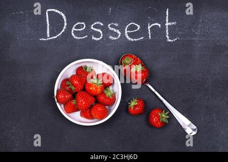 Un bol de fraises fraîches mûres avec une cuillère sur un tableau à craie, comme dessert du jour. Banque D'Images