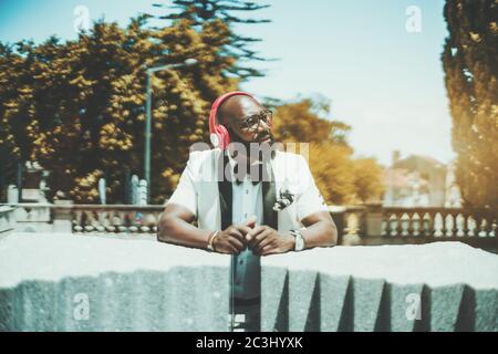 Beau homme africain à tête blanche et barbu en lunettes et élégant costume blanc est de profiter du temps ensoleillé d'été, debout à l'extérieur, et écouter de la musique Banque D'Images