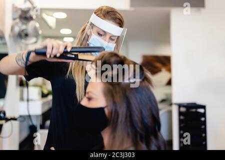 Coiffeur, protégé par un masque, peignant les cheveux de son client avec un fer à repasser dans un salon. Banque D'Images